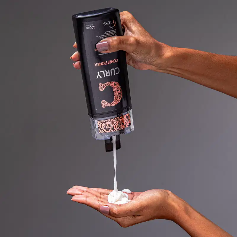 A imagem mostra uma mão segurando um frasco de condicionador da marca Truss, linha Curly. Também é possível ver o seu conteúdo, que é cremoso e branco, com 300ml.