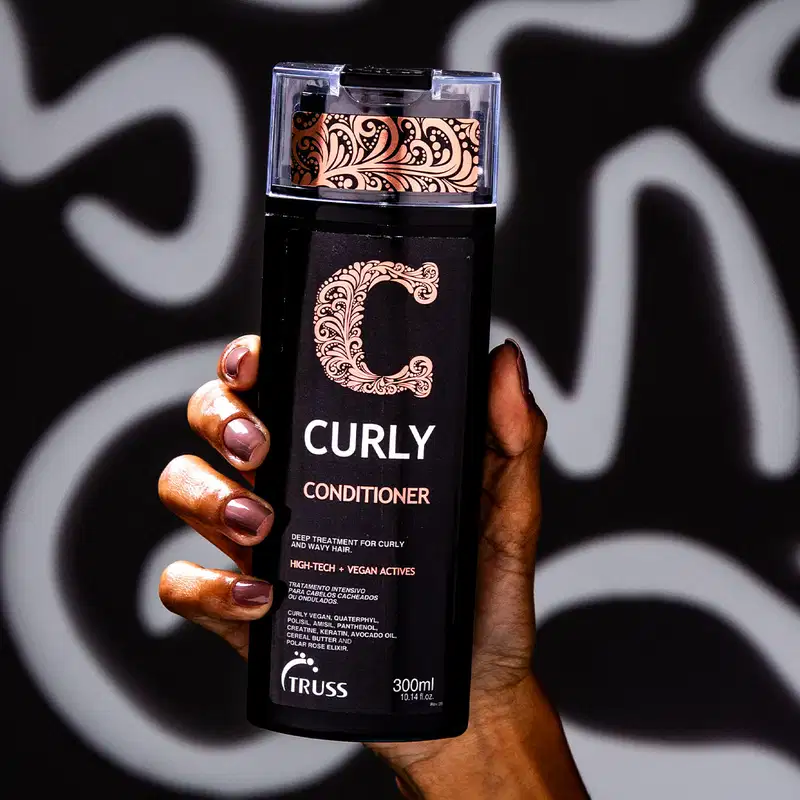 A imagem mostra uma mão segurando um frasco de condicionador da marca Truss, linha Curly. Também é possível ver o seu conteúdo, que é cremoso e branco, com 300ml.