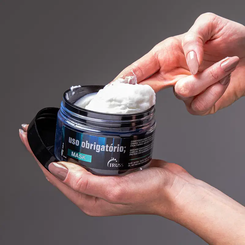 A imagem mostra uma mão está segurando o pote da máscara capilar e aplicando o produto branco cremoso com os dedos.