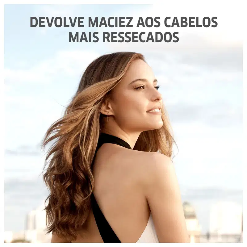 A imagem apresenta uma mulher jovem com longos cabelos ondulados em tons de loiro e castanho. Ela está usando um top preto e tem um sorriso suave no rosto. O texto diz "Devolve maciez aos cabelos mais ressecados".