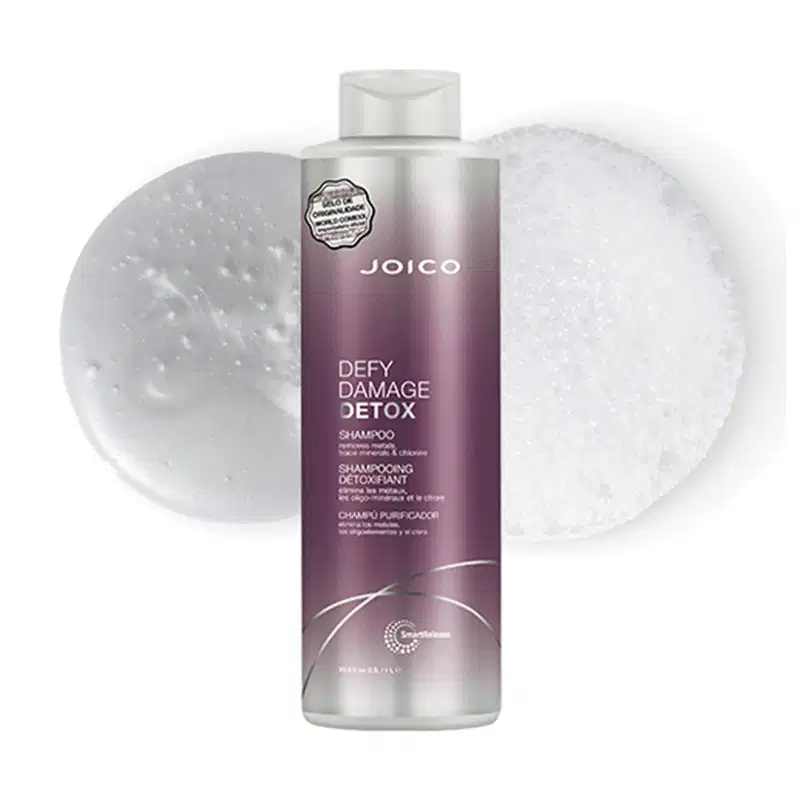 A imagem mostra o Shampoo Joico Defy Damage Detox e no fundo uma amostra com a textura e cor do produto, em tonalidade branca e espuma cremosa.