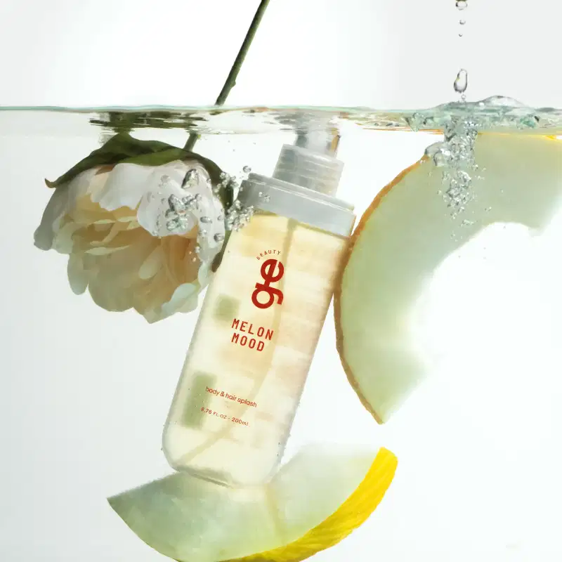 Frasco de body & hair splash Melon Mood da GE Beauty, 200ml, com líquido translúcido amarelado. Embalagem branca com rótulo vermelho. Apresentado em composição com melão fresco e flor branca em água.