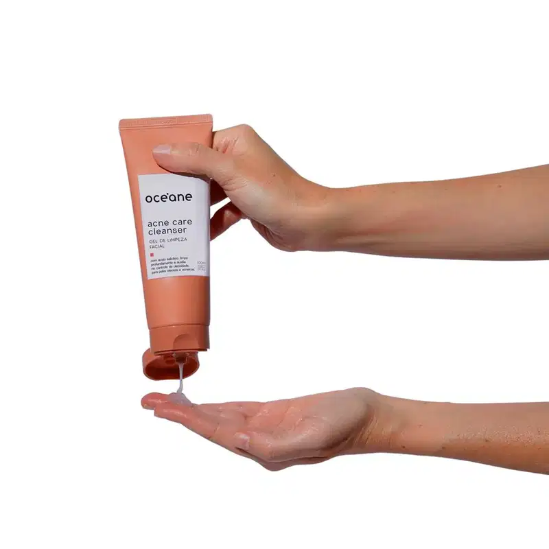 A imagem mostra um par de mãos segurando a embalagem do Océane Acne Care Cleanser. Uma das mãos está apertando o tubo para dispensar o produto líquido na outra mão aberta.