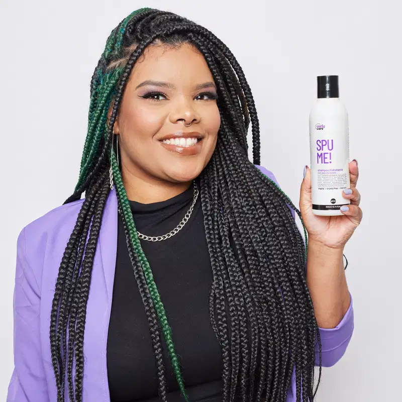 O produto mostrado é um shampoo chamado "Spu Me!". O O frasco está sendo segurado por uma pessoa com longos cabelos negros trançados, que incluem detalhes em verde