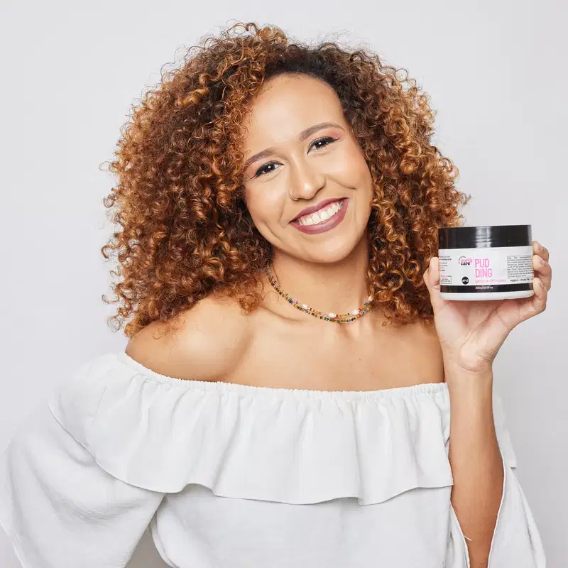 Pote plástico branco com tampa preta contendo um produto de definição de cachos da marca Curly Care. O rótulo indica que é um "Pudding Gelatina Definidora" vegano, com pH 4,2.