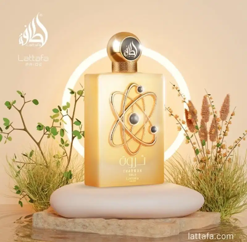 Um item de perfumaria dourado com um símbolo atômico em destaque, posicionado sobre uma superfície de madeira. Ao redor, há plantas verdes e flores secas, criando um ambiente natural e elegante.