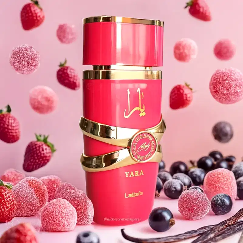 Ambiente de fragrância Lattafa Perfumes Yara, com embalagem vermelha e dourada, cercada por frutas vermelhas e azuis, em um cenário rosa, sugerindo uma ocasião sofisticada e elegante.