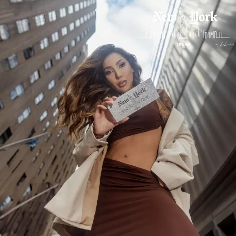 A imagem mostra uma mulher posando em um ambiente urbano com edifícios ao fundo. Ela está segurando a paleta de sombras. A mulher tem cabelos longos e ondulados, está usando um casaco branco aberto que revela sua barriga nua.