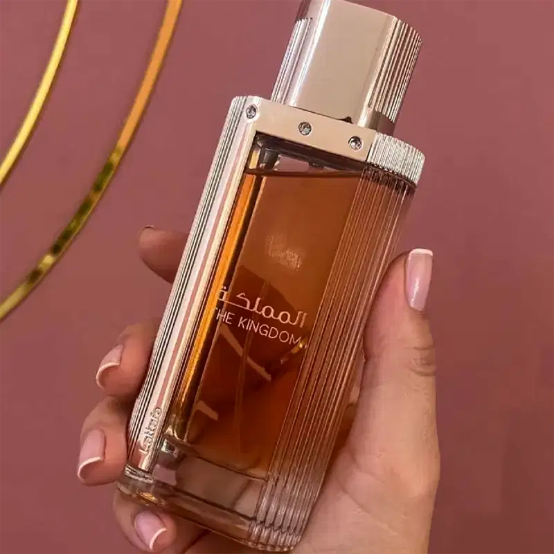 A imagem mostra um frasco de perfume da marca Lattafa Perfumes, com o nome "The Kingdom" impresso no vidro. O produto é segurado por uma mão feminina com unhas pintadas, em um ambiente com fundo rosa, sugerindo um contexto de uso pessoal e elegante.