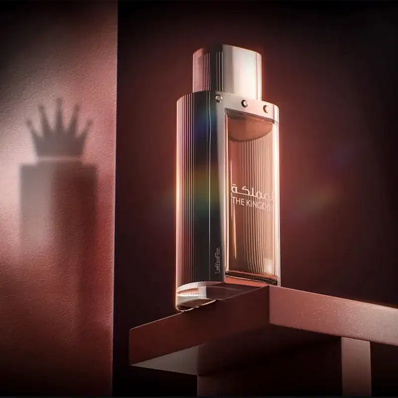 Ambiente escuro e elegante, com iluminação suave, destacando um frasco de perfume da marca Lattafa Perfumes, intitulado "The Kingdom". O produto é o foco central, sugerindo um contexto de luxo e sofisticação.
