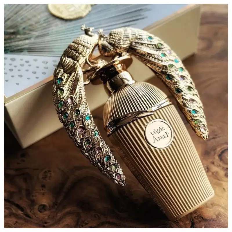 Frasco de perfume Lattafa Affee com embalagem decorativa em metal dourado, adornada com pedras coloridas em formato de penas. O rótulo do produto é visível na parte frontal do frasco.