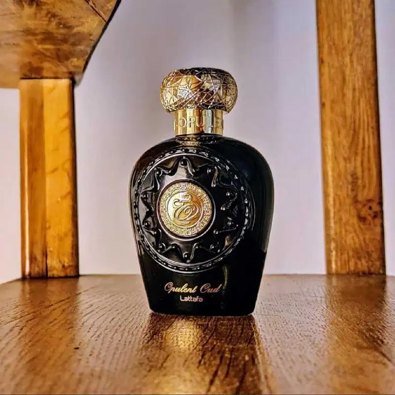 Ambiente elegante e sofisticado, com detalhes dourados e pretos que realçam a embalagem de perfume da marca Lattafa. O produto é apresentado em um cenário de madeira, sugerindo um contexto de luxo e exclusividade.