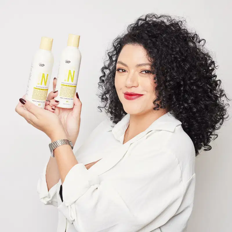 A imagem mostra dois frascos de produtos para cabelos cacheados da marca Curly Care. Os frascos são brancos e alongados, com tampas amarelas. Os frascos estão sendo segurados por uma mulher de cabelos cacheados escuros e volumosos.
