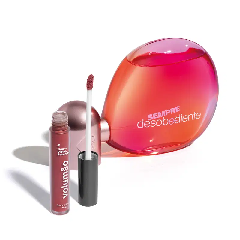 Imagem do Eau de Parfum Sempre Desobediente com vidro transparente em formato oval, líquido rosa e letras brancas e batom líquido Volumão Roseli na cor rosa em embalagem transparente e aplicador macio também marrom. O fundo é branco com sombreado.