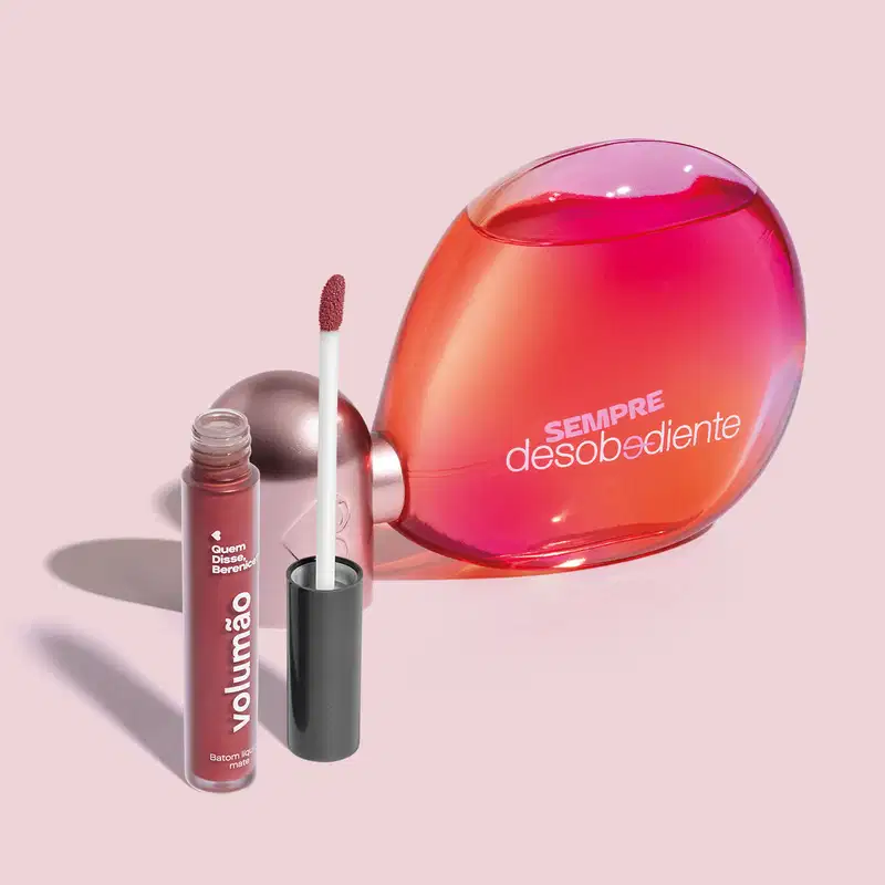 Imagem do Eau de Parfum Sempre Desobediente com vidro transparente em formato oval, líquido rosa e letras brancas e batom líquido Volumão Roseli na cor rosa em embalagem transparente e aplicador macio também marrom. O fundo é bege com sombreado.