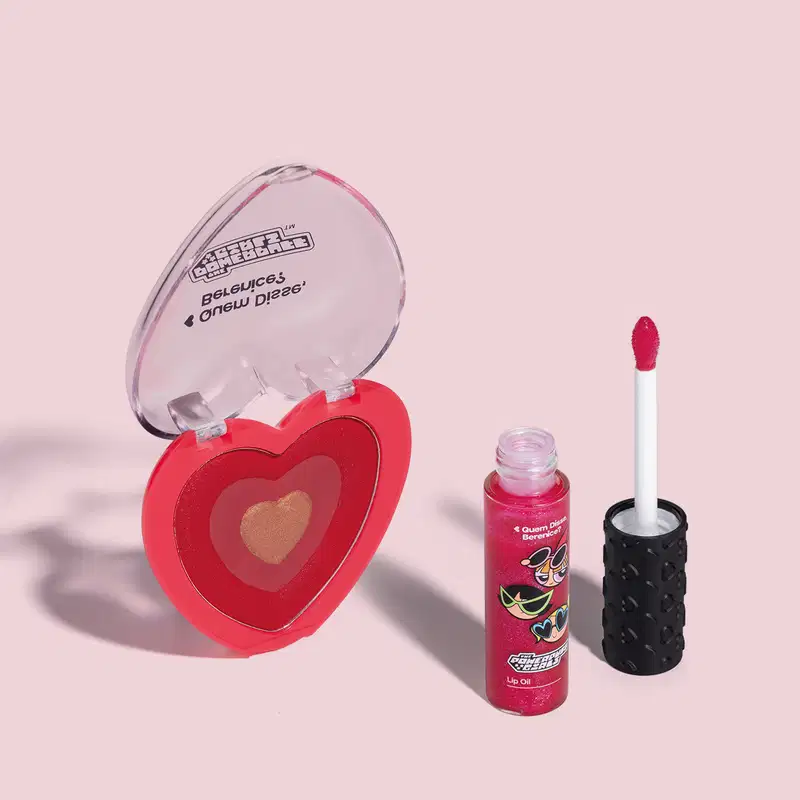 Dois itens da marca Meninas Superpoderosas Quem Disse, Berenice?: um lip oil vermelho em embalagem transparente com personagens e um blush compacto em formato de coração vermelho com tampa transparente em fundo lilás com leve sombreado.