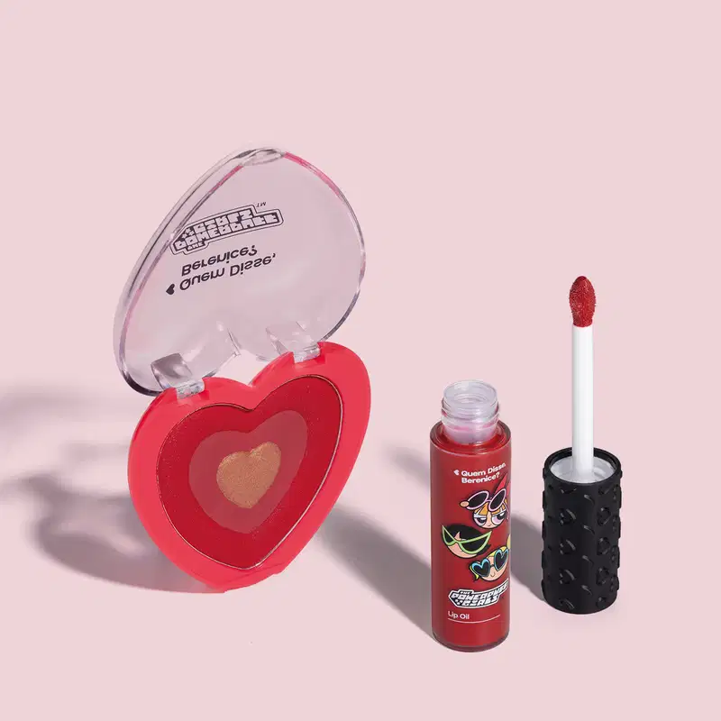 Imagem de dois itens da marca QDB As Meninas Super Poderosas, sendo um blush compacto rosa em formato de coração e um lip oil vermelho com aplicador em forma de esponja na cor vermelha. O fundo da imagem é neutro com sombreado.