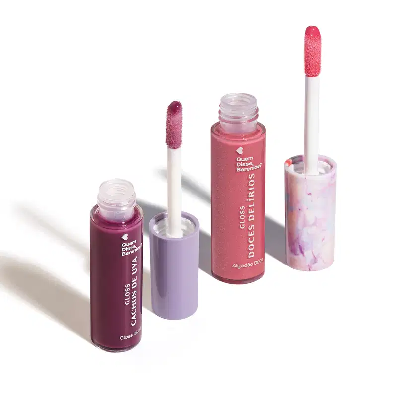Imagem de dois frascos de gloss labial, um roxo com título Cachos de Uva e outro rosa com título Doces Delírios. Ambos em recipiente transparente com aplicador em forma de esponja na mesma cor do gloss. O fundo da imagem é branco com sombreado.