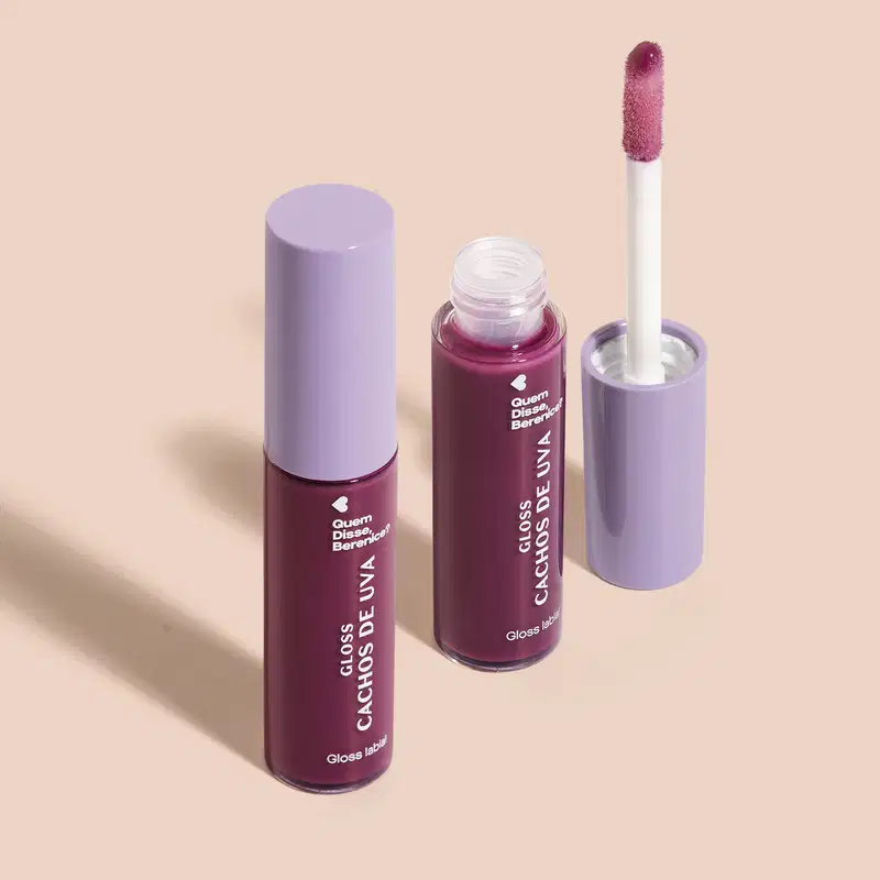 Imagem de combo com dois frascos de 10ml contendo gloss labial Cachos de Uva QDB E Cuide-se Bem Quem Disse, Berenice?. A embalagem é transparente com conteúdo roxo, tampa cinza e aplicador macio na cor roxa. O fundo é bege com leve sombreado.
