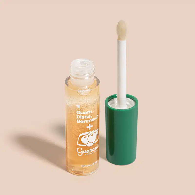 Frasco com líquido dourado e rótulo com texto em português, acompanhado de um aplicador de maquiagem branco em um suporte verde. O produto é identificado como "Quem disse, Berenice?" e "Gloss Labial", indicando tratar-se de um gloss labial.