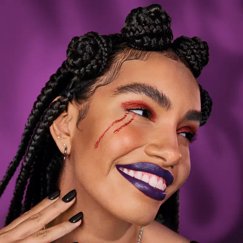 Imagem de uma mulher usando tranças finas no cabelo escuro, esmalte preto e maquiagem colorida típica de Halloween, trazendo o glitter usado como lágrimas. Ela sorri. O fundo é roxo vibrante.