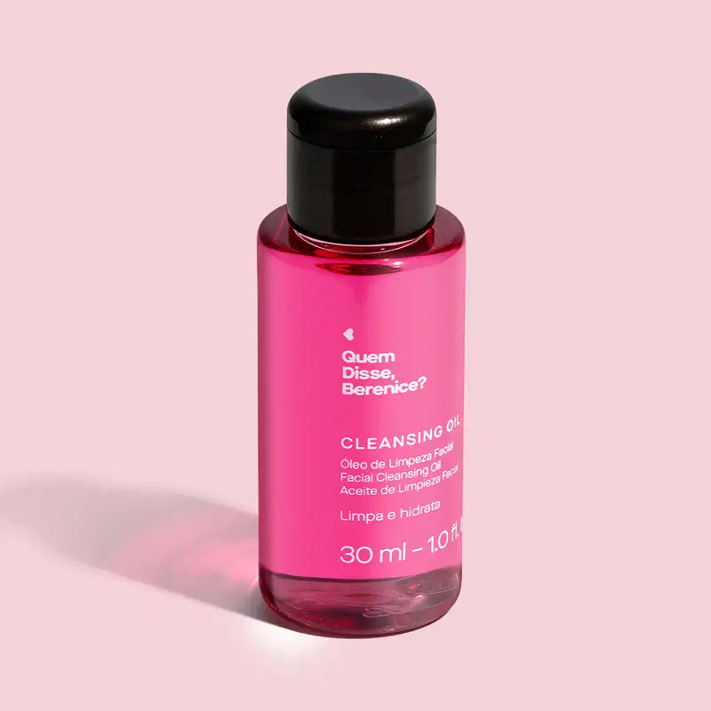 Imagem de frasco transparente e tampa preta contendo um líquido rosa com rótulo branco "Quem disse, Berenice? Cleansing Oil" em preto, trazendo 30 ml de óleo de limpeza facial. Ele está em fundo em tom de rosa bem claro e um leve sombreado da embalagem.