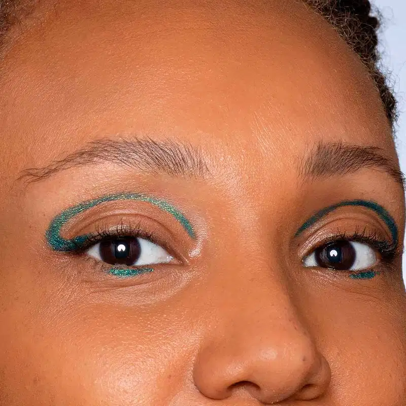 Olhos de uma pessoa com maquiagem colorida em tons de marrom e verde. A imagem mostra os olhos de perto, com sobrancelhas bem definidas e cílios destacados. A maquiagem parece ser aplicada com cuidado e detalhe, criando um visual elegante e expressivo.