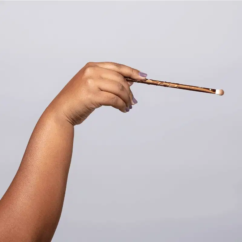 A imagem mostra uma mão segurando um pincel de maquiagem comprido e fino. O pincel tem um cabo de madeira com detalhes em tons de marrom e uma ponta fina com cerdas brancas. A mão é de pele parda e tem uma unha pintada com esmalte lilás claro.