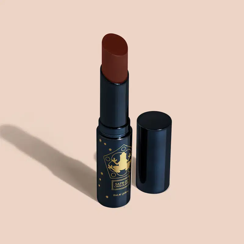Balm em formato de bastão com uma cor marrom. A embalagem é um tubo azul escuro com detalhes dourados que incluem um símbolo de um caldeirão. O produto tem uma aparência elegante e sofisticada, com o tubo azul fosco contrastando com os detalhes dourados