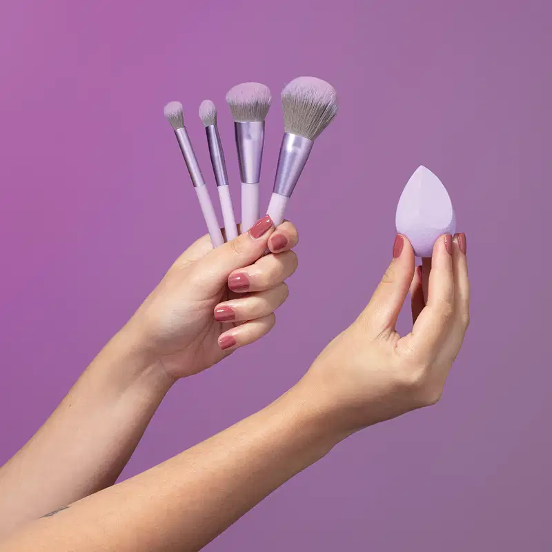 Duas mãos femininas com unhas pintadas, uma delas segura quatro pinceis de maquiagem e a outra segura uma esponja de maquiagem em formato de gota em fundo roxo.