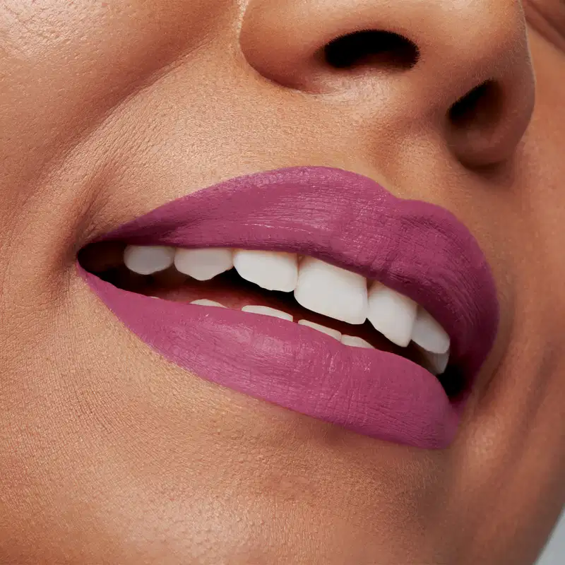 Close-up de um rosto feminino que está sorrindo, mostrando os dentes brancos e evidenciando os lábios que usam o batom líquido Volumão Roxo Ameixali Quem Disse, Berenice?.