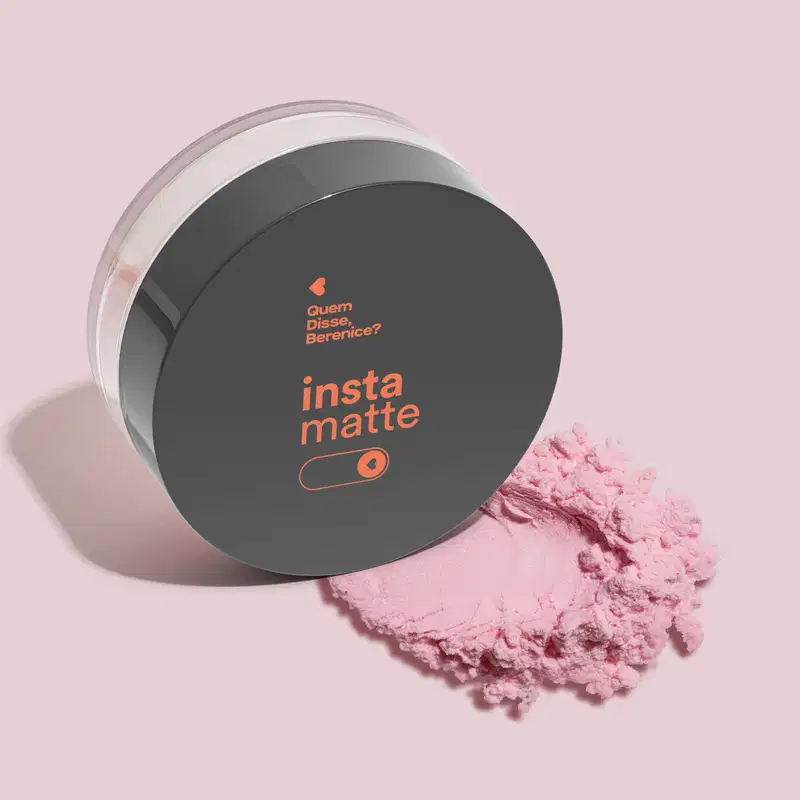 Pote redondo de acrílico transparente e tampa preta apoiado lateralmente sobre superfície branca como as marcas "Insta matte" e "Quem disse, Berenice?" impressas em cor laranja. Abaixo do pote, há um pouco de pó facial de cor rosa sobre superfície rosa.