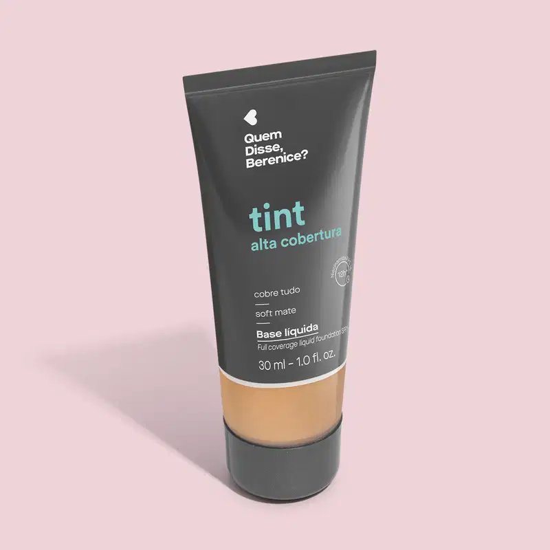 A imagem mostra a Base Tint Alta Cobertura da Quem Disse, Berenice? Embalagem cinza escura com parte inferior transparente para mostrar a cor do produto sobre fundo rosa.