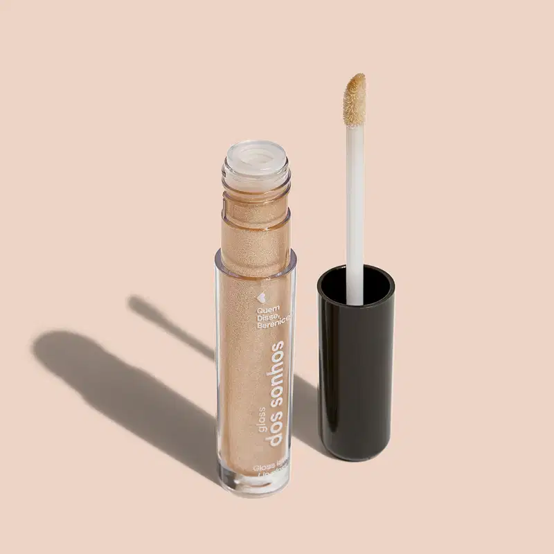 Frasco cilíndrico transparente contendo um brilho labial dourado brilhante. Rótulo na embalagem com o nome do produto "Gloss dos Sonhos" e a descrição "Quem disse, berence?". Ao lado, a tampa preta do produto, que acompanha um aplicador.
