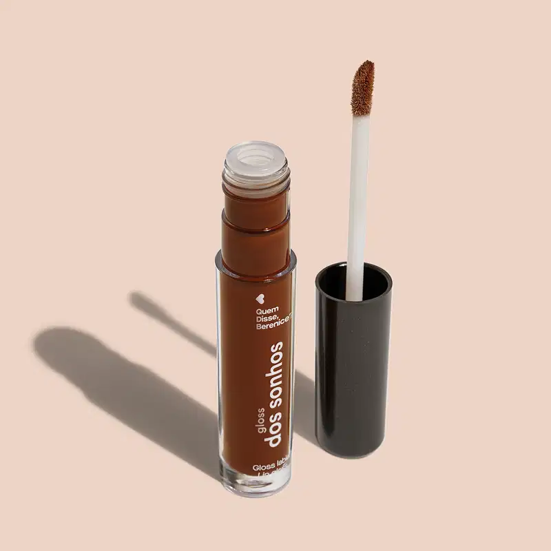 Frasco cilíndrico transparente contendo um gloss labial marrom. Rótulo na embalagem com o nome do produto "Gloss dos Sonhos" e a descrição "Quem disse, berence?". Ao lado, a tampa preta do produto, que acompanha um aplicador de espuma.