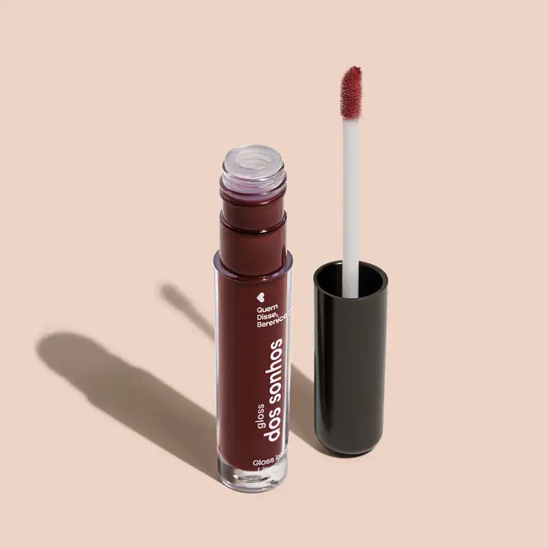 Frasco cilíndrico transparente contendo um gloss labial roxo. Rótulo na embalagem com o nome do produto "Gloss dos Sonhos" e a descrição "Quem disse, berence?". Ao lado, a tampa preta do produto, que acompanha um aplicador de espuma.