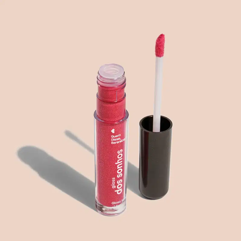 Frasco cilíndrico transparente contendo um gloss labial rosa brilhante Rótulo na embalagem com o nome do produto "Gloss dos Sonhos" e a descrição "Quem disse, berence?". Ao lado, a tampa preta do produto, que acompanha um aplicador de espuma rosa.
