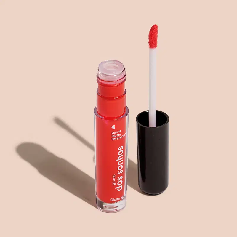 Frasco cilíndrico transparente contendo um gloss labial vermelho. Rótulo na embalagem com o nome do produto "Gloss dos Sonhos" e a descrição "Quem disse, berence?". Ao lado, a tampa preta do produto, que acompanha um aplicador de espuma.