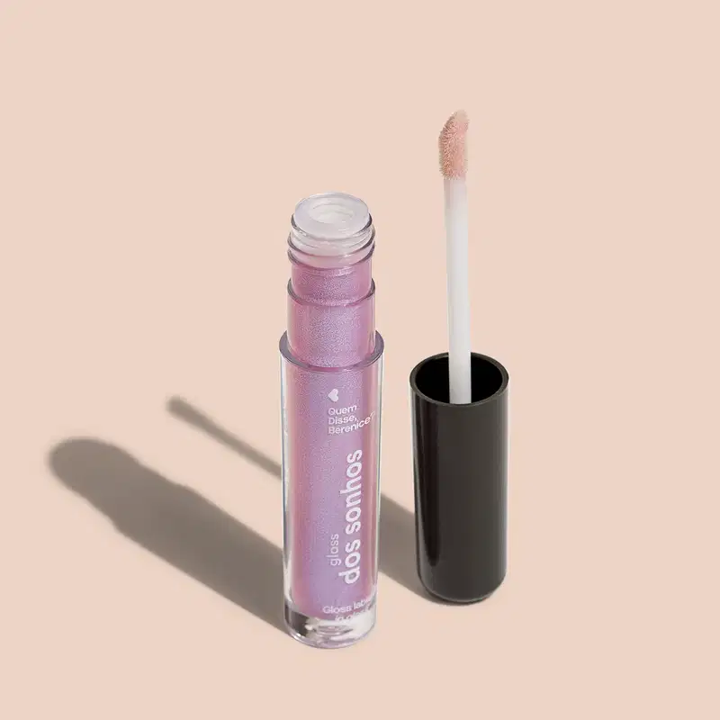 Frasco cilíndrico transparente contendo um gloss labial rosa com púrpura. Rótulo na embalagem com o nome do produto "Gloss dos Sonhos" e a descrição "Quem disse, berenice?". Ao lado, a tampa preta do produto, que acompanha um aplicador de espuma.