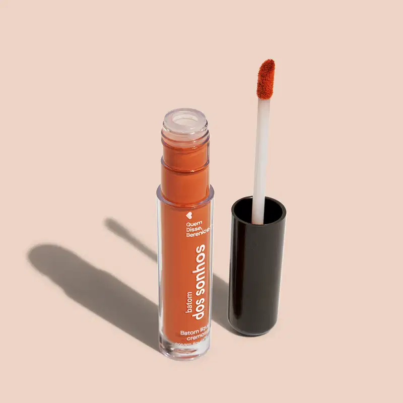 Batom líquido Laranja em frasco transparente de plástico, com aplicador de espuma. O frasco possui uma tampa protetora preta para guardar o aplicador. O rótulo no frasco exibe o nome da marca e do produto em letras brancas sobre fundo laranja.