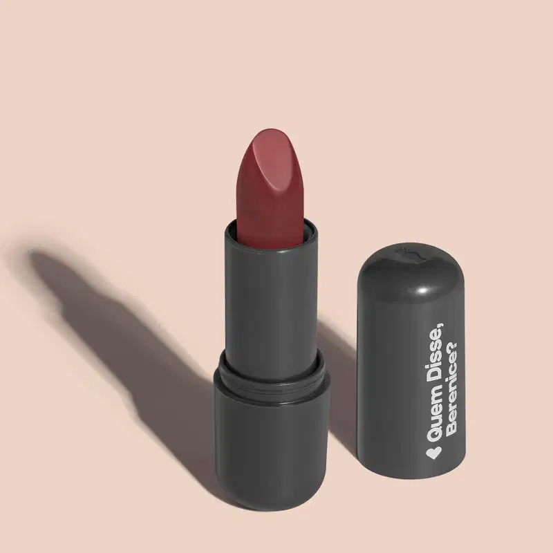 Batom de cor vermelho escuro em embalagem preta com logotipo da marca Quem Disse, Berenice? em destaque. O batom está aberto sobre superfície rosa com iluminação que cria uma longa sombra projetada atrás da embalagem.