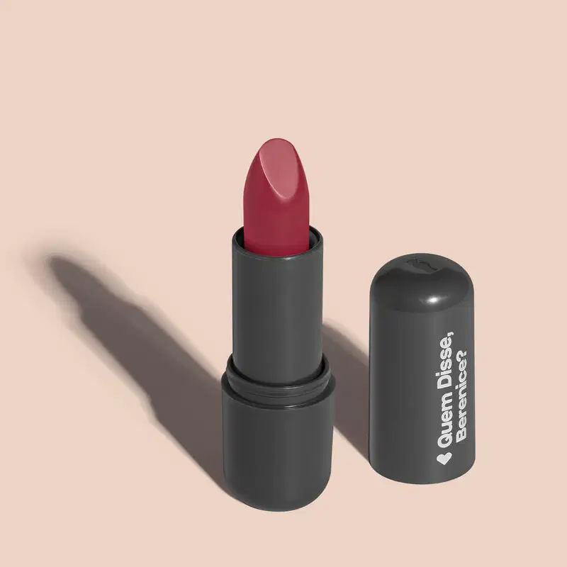 Batom de cor rosa avermelhado em embalagem preta com logotipo da marca Quem Disse, Berenice? em destaque. O batom está aberto sobre superfície rosa com iluminação que cria uma longa sombra projetada atrás da embalagem.