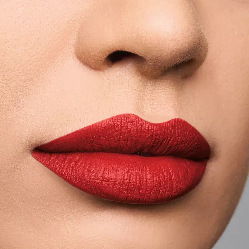 A imagem mostra um close-up de lábios com uma aplicação de batom vermelho intenso. O batom possui uma textura cremosa e brilhante, conferindo um acabamento aveludado aos lábios. A cor do batom é um vermelho vibrante,.