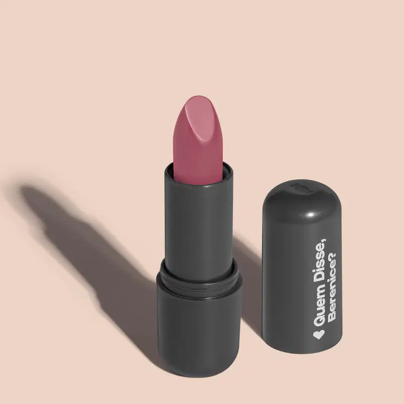 Batom de cor rosa claro em embalagem preta com logotipo da marca Quem Disse, Berenice? em destaque. O batom está aberto sobre superfície rosa com iluminação que cria uma longa sombra projetada atrás da embalagem.