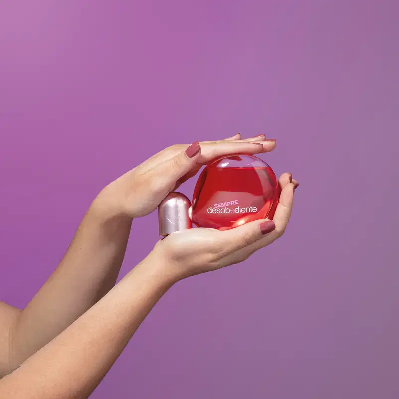 Recipiente vermelho e rosa de Eau de Parfum Desobediente Quem Disse, Berenice? em mãos femininas sobre fundo roxo.