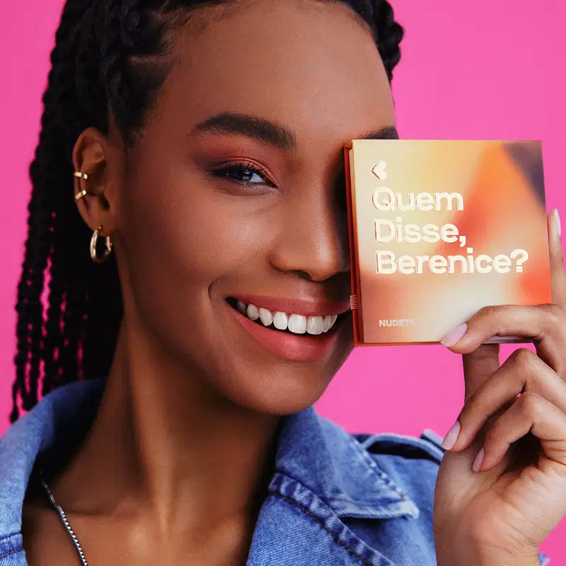 O produto mostrado na imagem é uma paleta de maquiagem o título "Quem disse, Berenice?" em letras brancas. A pessoa na imagem segura o livro e sorri, com um fundo rosa vibrante.