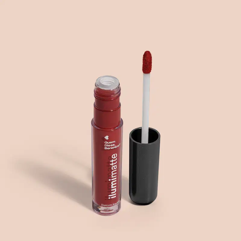 Frasco de batom líquido vermelho Vermelumi Ilumimatte Quem Disse, Berenice? aberto, mostrando a tonalidade do produto,, com a tampa preta ao lado que traz haste branca e esponja aplicadora na cor marrom em fundo rosado com leve sombreado.