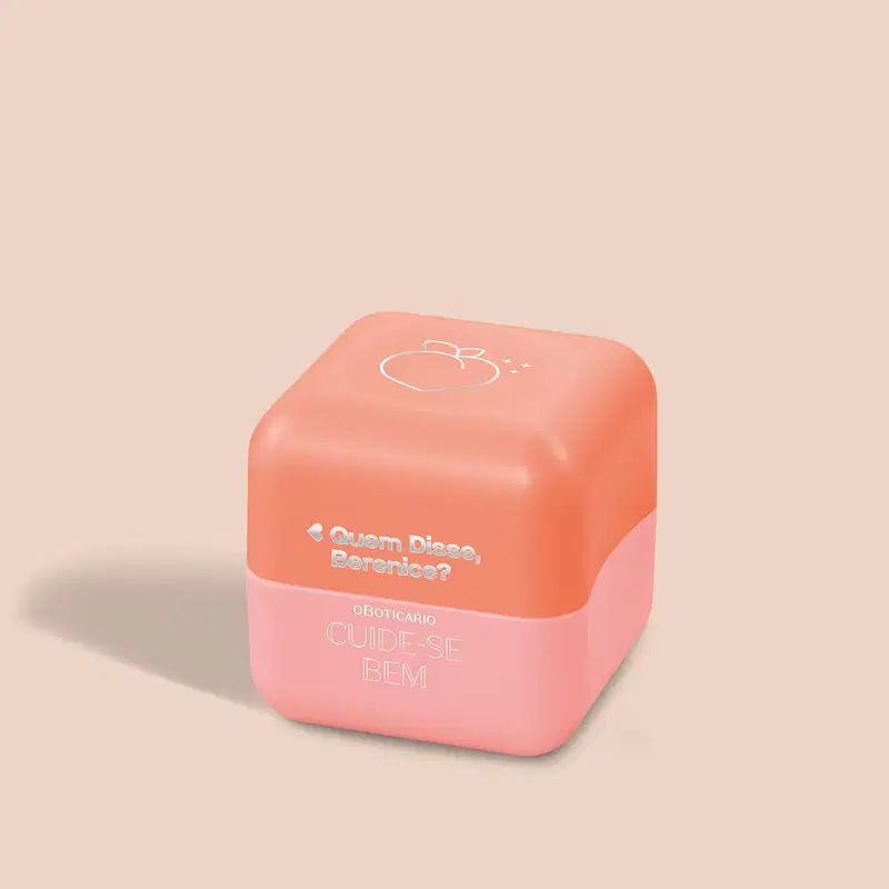 Balm labial em embalagem plástica hexagonal na cor coral. A parte superior da embalagem é mais escura, com um desenho estilizado de um pêssego. Na parte inferior, texto em letras brancas: "Quem Disse, Berenice? OBOTICÁRIO CUIDE-SE BEM".