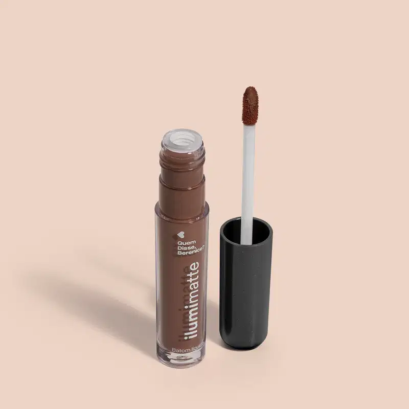 Frasco de batom líquido marrom Marronlumi Ilumimatte Quem Disse, Berenice? aberto, mostrando a tonalidade do produto,, com a tampa preta ao lado que traz haste branca e esponja aplicadora na cor marrom em fundo rosado com leve sombreado.