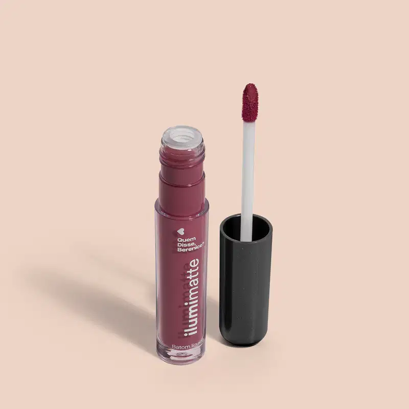 Frasco de batom líquido malva Malvalumi Ilumimatte Quem Disse, Berenice? aberto, mostrando a tonalidade do produto,, com a tampa preta ao lado que traz haste branca e esponja aplicadora na cor rosa escuro em fundo rosado com leve sombreado.